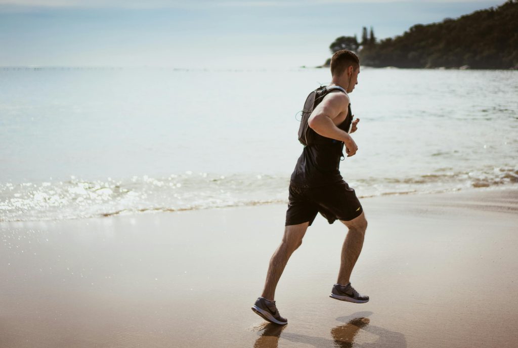 "Homem correndo na praia com mochila de hidratação, representando estilo de vida saudável e prática esportiva — ideal para ilustrar artigo com dicas para iniciantes de corrida."
